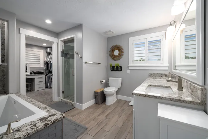 Wood-finish flooring, dual vanity with undermount sinks and stone countertops, glass-enclosed shower, built-in soaking tub, and window with plantation shutters