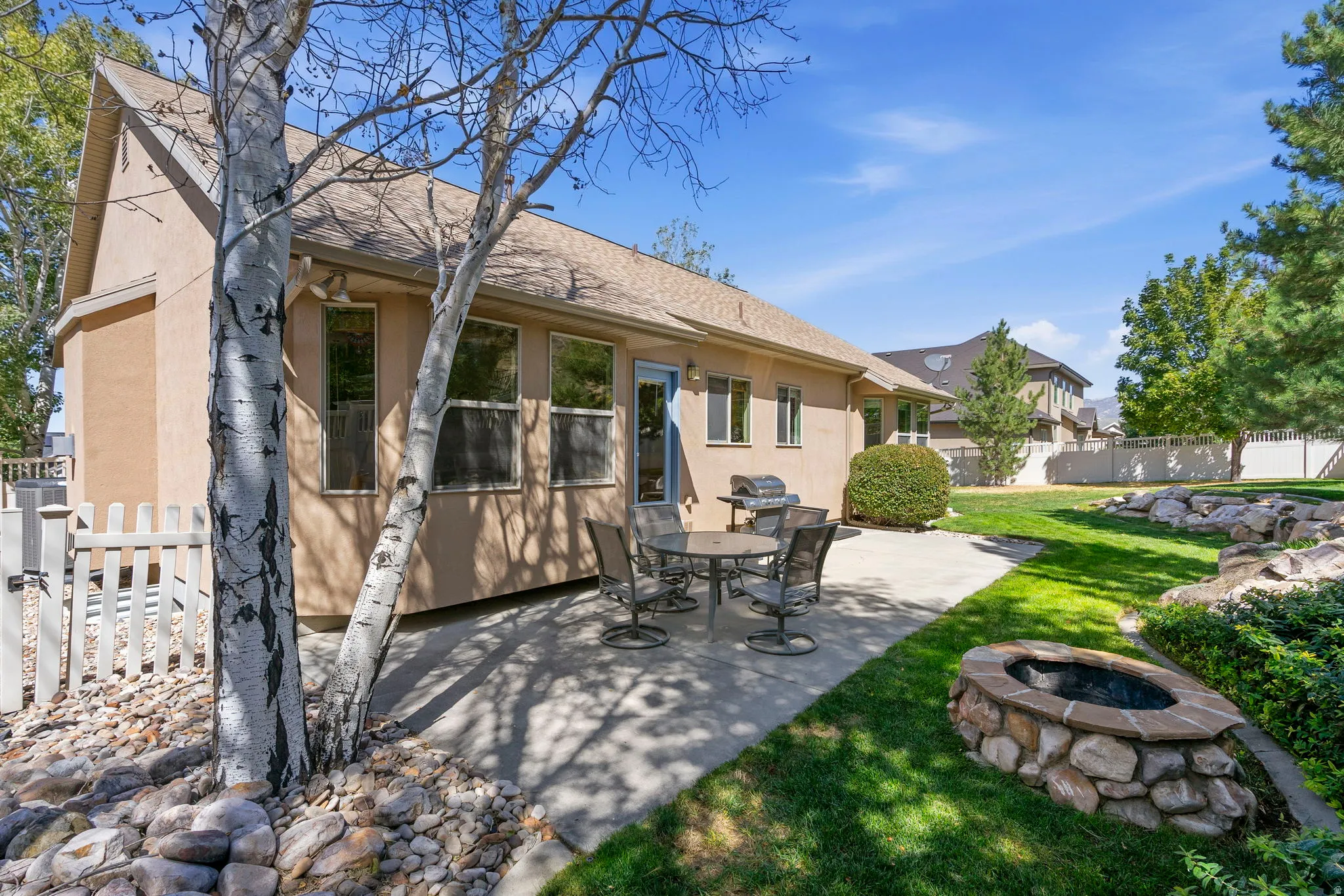 Expansive outdoor patio with a built-in stone fire pit, mature landscaping, a manicured lawn, and a light-toned stucco exterior