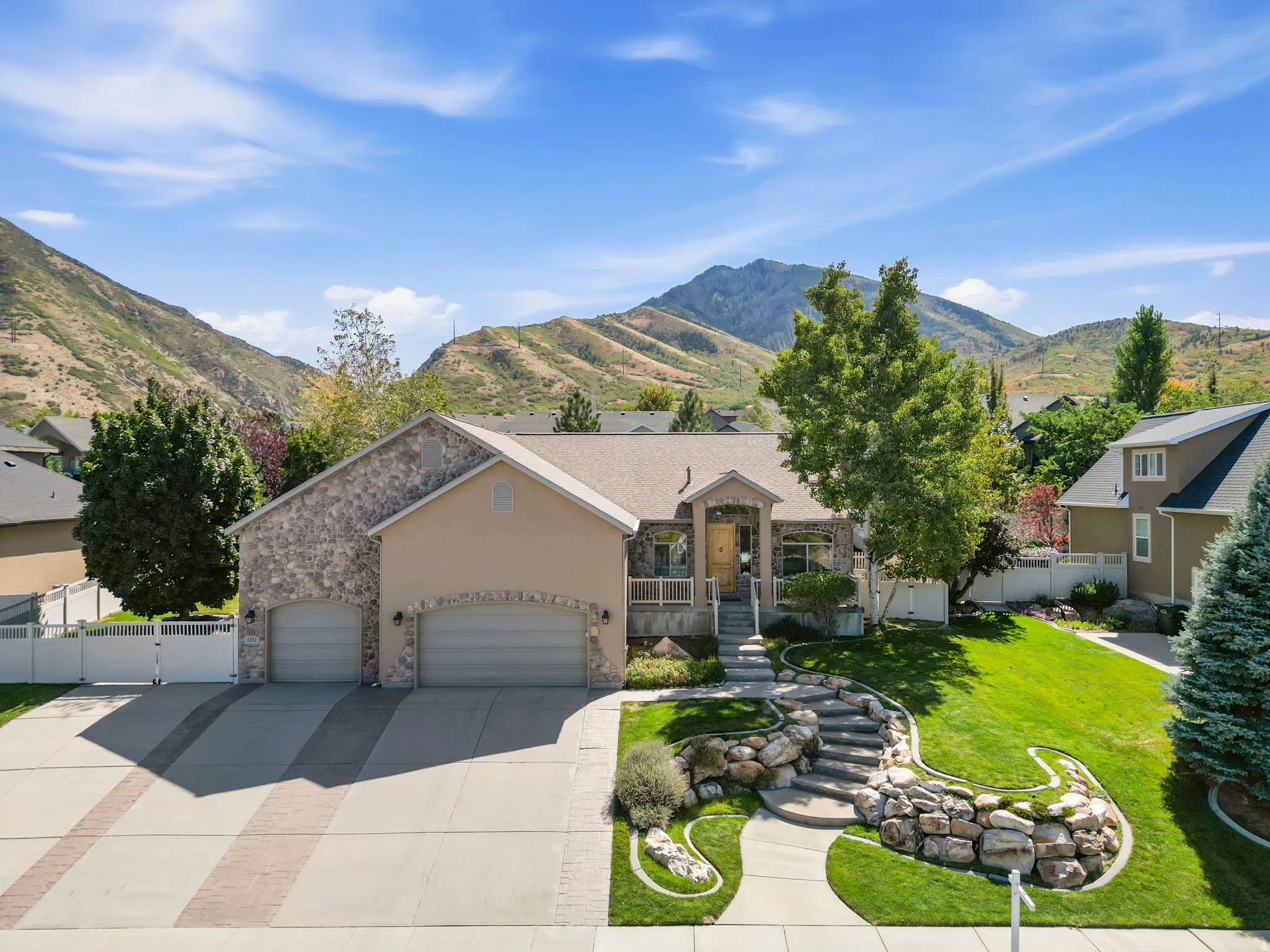 Stone facade home with an integrated two-car garage, a concrete driveway featuring brick accents, and a landscaped front yard with rock retaining walls and a paved walkway leading to the front entry