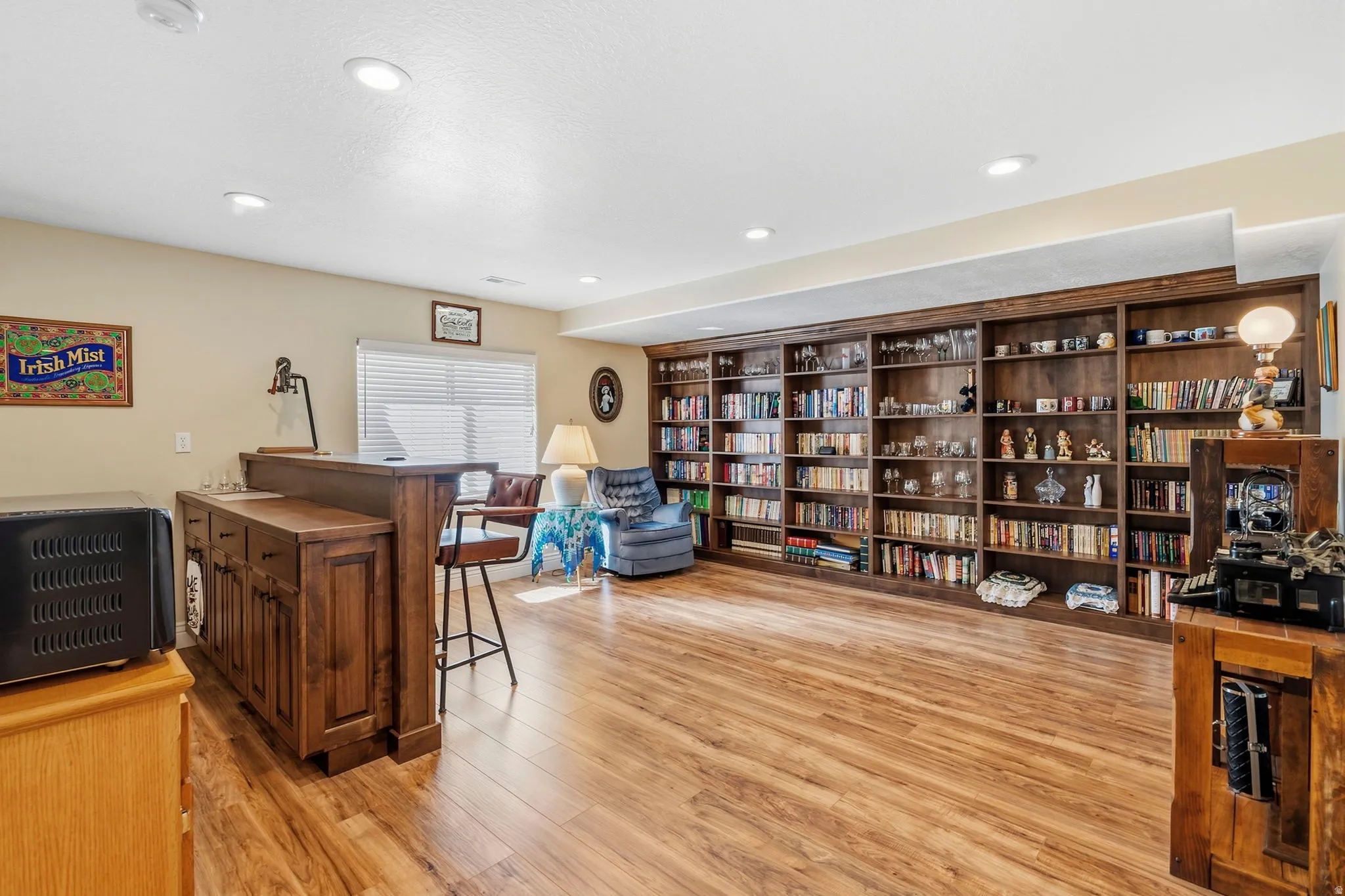 Built-in wood-finish shelving, recessed ceiling lighting, wood-finish flooring, built-in wet bar with wood-finish cabinetry, and a window with horizontal blinds