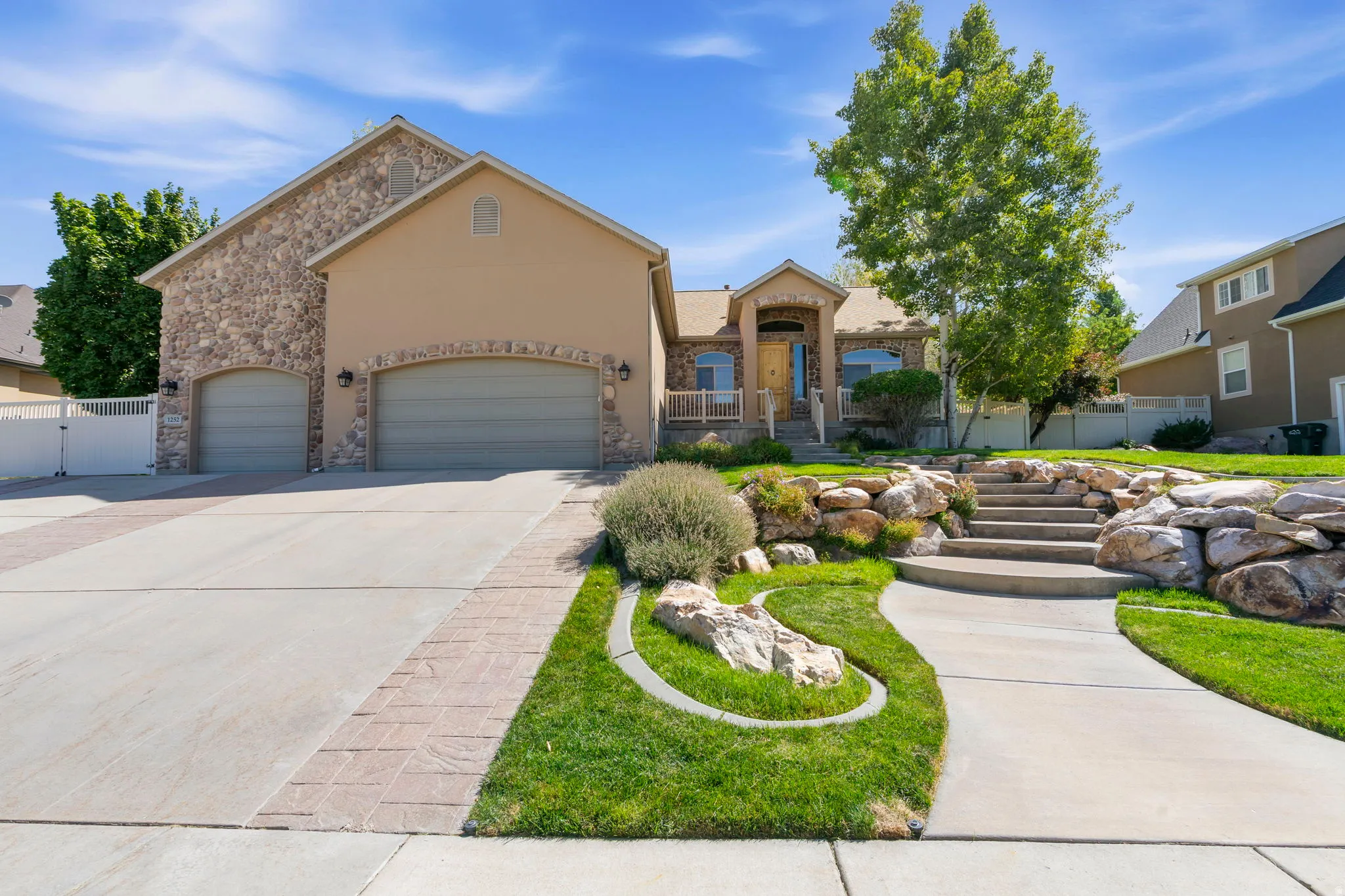 Stone and stucco exterior, arched garage doors, concrete driveway with paver accents, tiered landscaping with rock retaining walls, and a concrete pathway leading to the entry