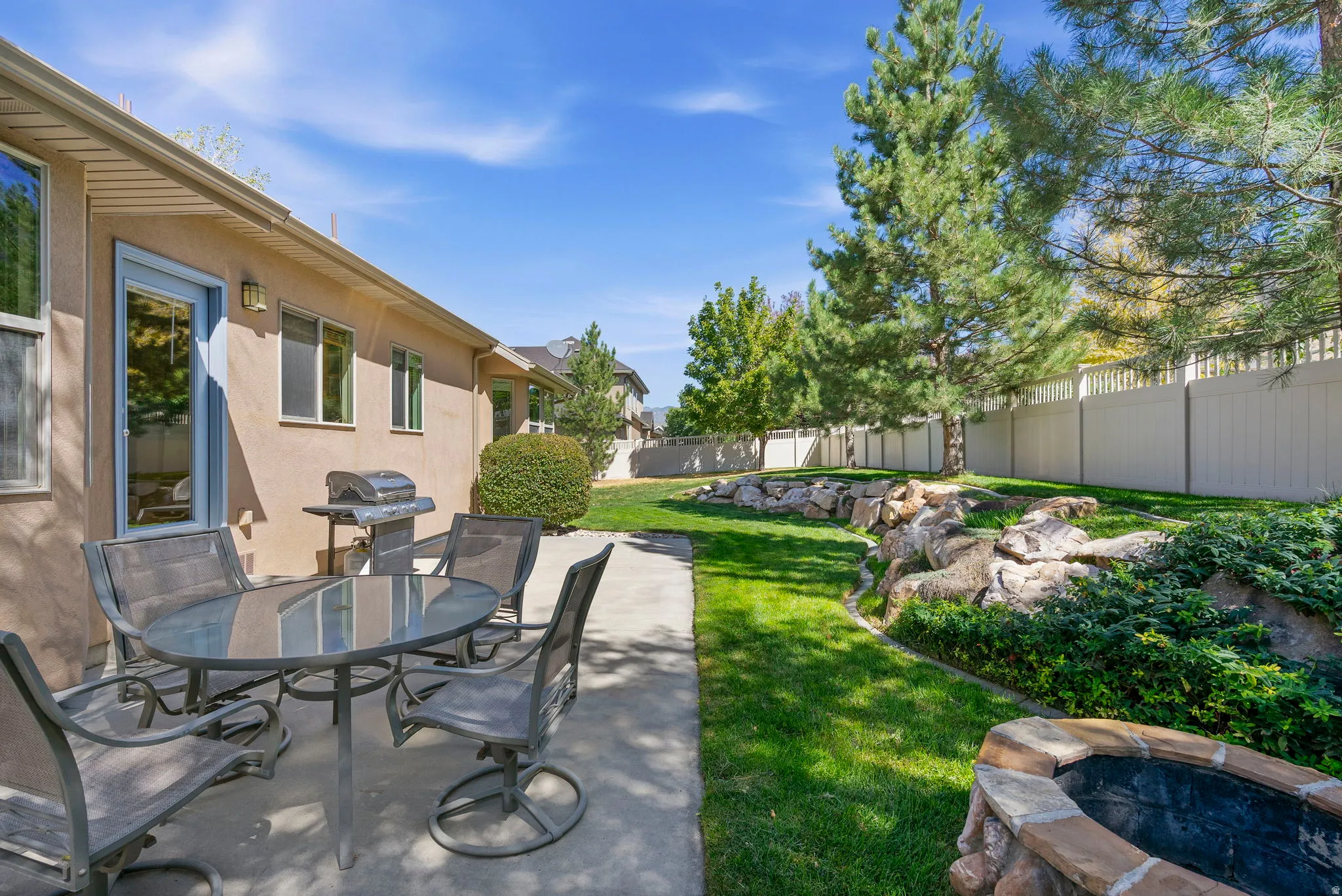 Expansive backyard featuring a concrete patio, a built-in stone-finish fire pit, mature trees, a rock feature with integrated plantings, and a privacy fence