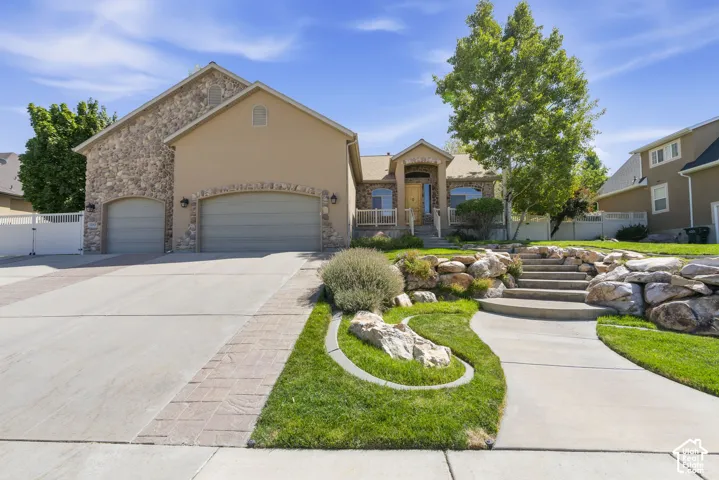 Stone and stucco exterior, arched garage doors, concrete driveway with paver accents, tiered landscaping with rock retaining walls, and a concrete pathway leading to the entry