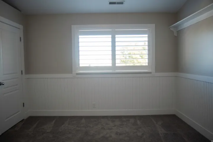 Window with white plantation shutters, white beadboard wainscoting, neutral wall paint, built-in shelving, and carpeted flooring
