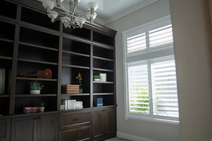 Built-in dark wood shelving unit with lower cabinetry and drawers, crown molding, and a multi-tiered window with plantation shutters