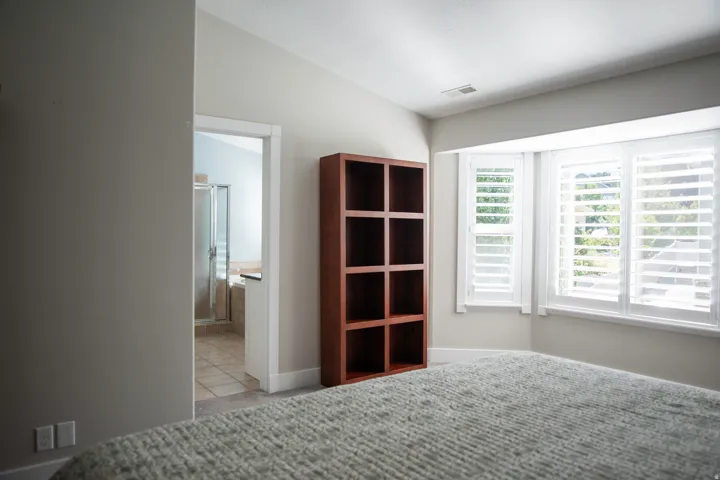 Recessed window with plantation shutters, wood-finish shelving unit, carpeted flooring, and a tiled bathroom with a glass-enclosed shower and soaking tub