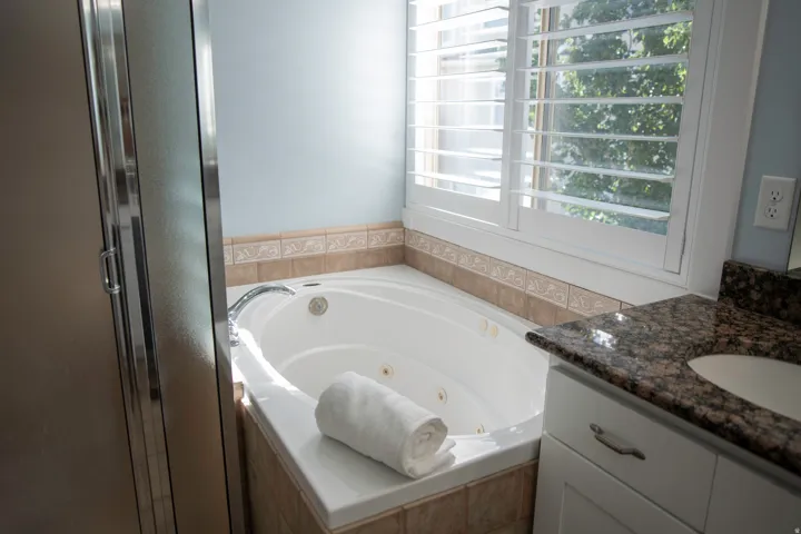 Corner jetted tub with tiled surround, white plantation shutters, built-in vanity with stone countertop, and glass-enclosed shower