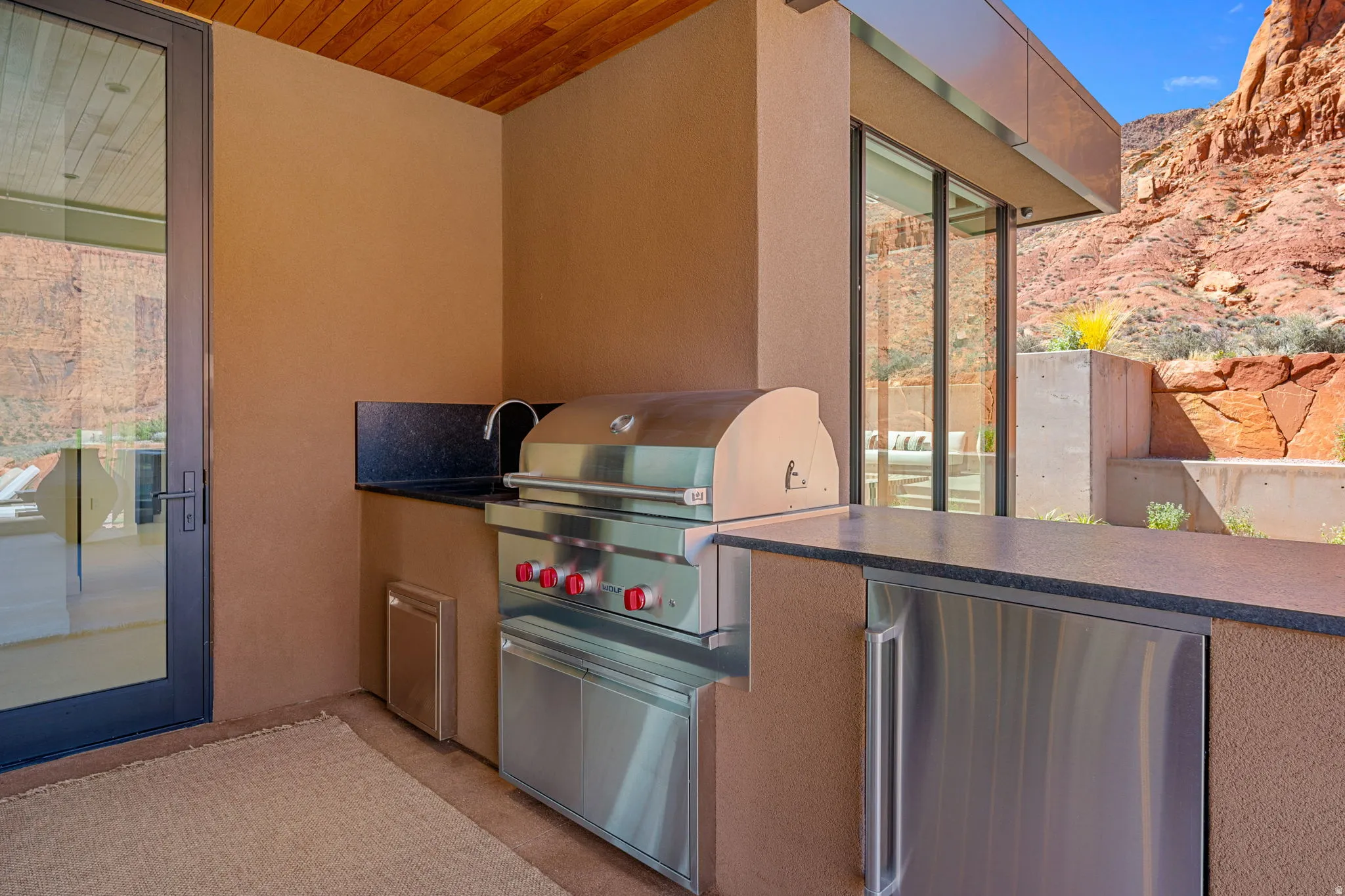 Outdoor kitchen featuring a built-in stainless steel grill, integrated sink, under-counter refrigerator, dark stone countertops, and a wood-finish ceiling