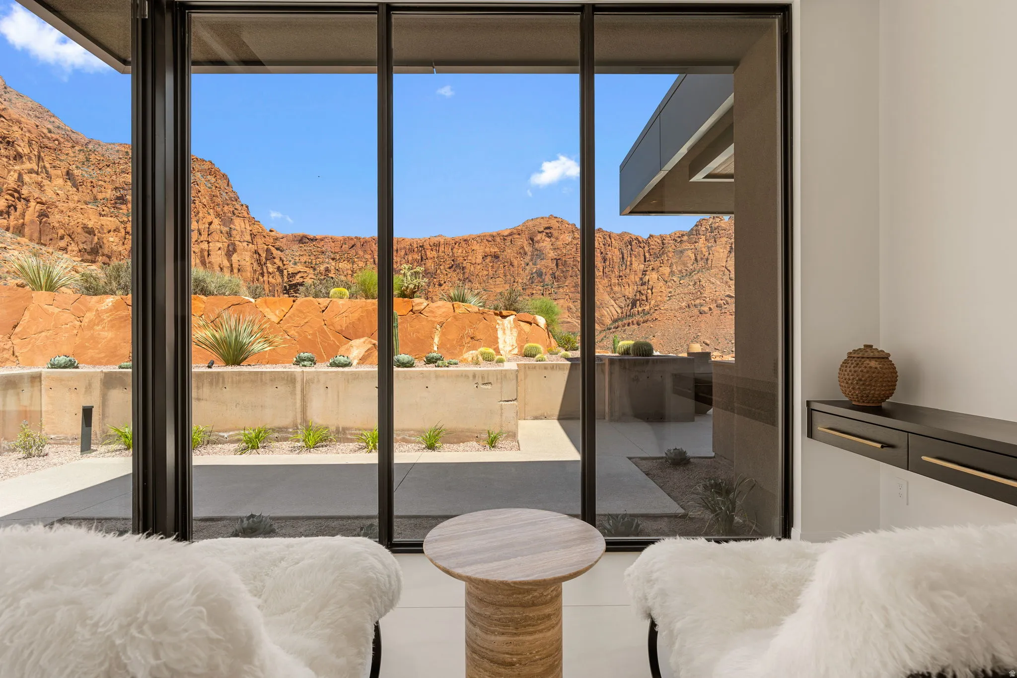 Floor-to-ceiling glass panel doors, mountain backdrop, exterior landscaping with desert flora, concrete retaining wall, and a built-in floating console