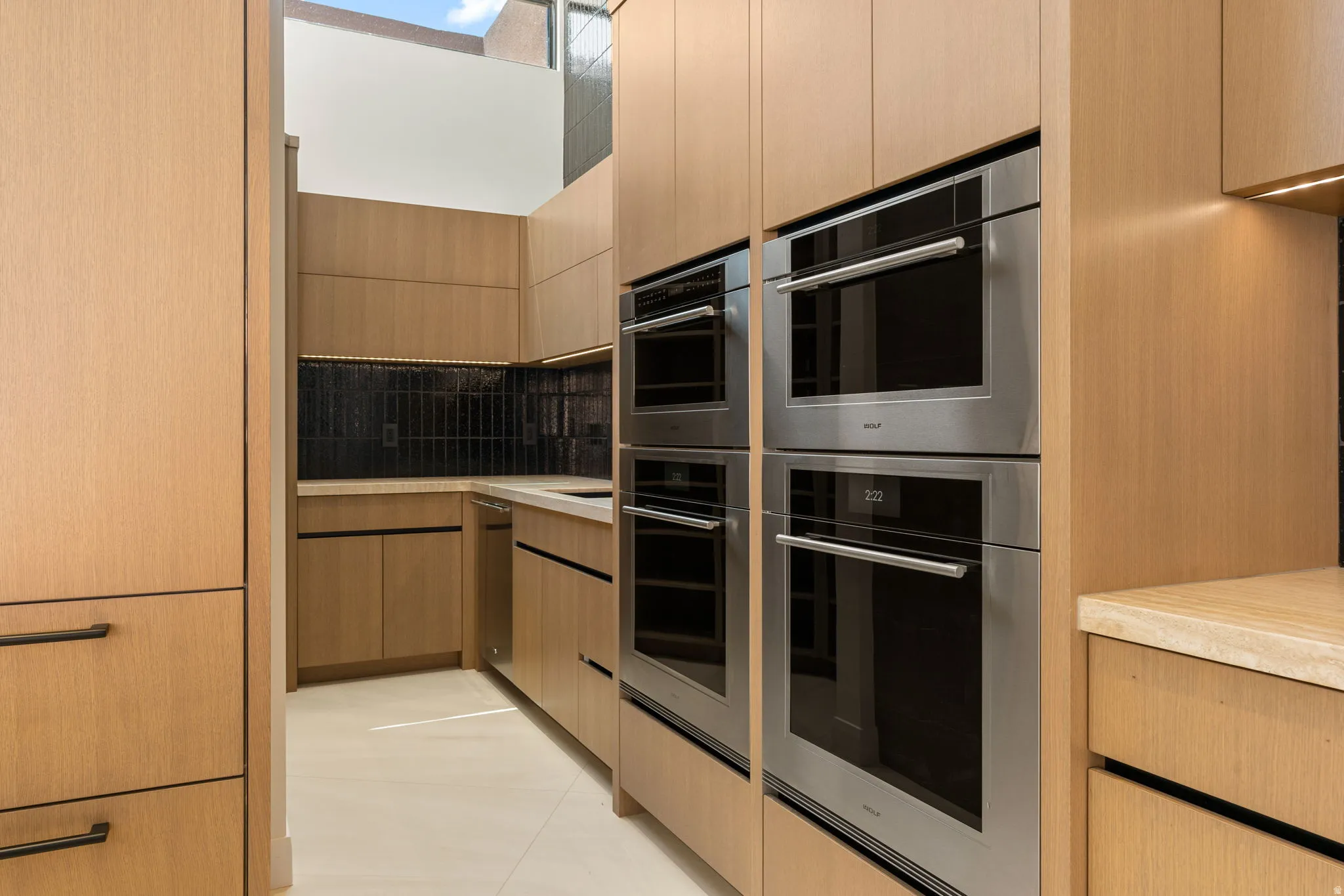 Light wood-finish cabinetry, integrated stainless steel appliances, dark tile backsplash, light countertops, and under-cabinet lighting