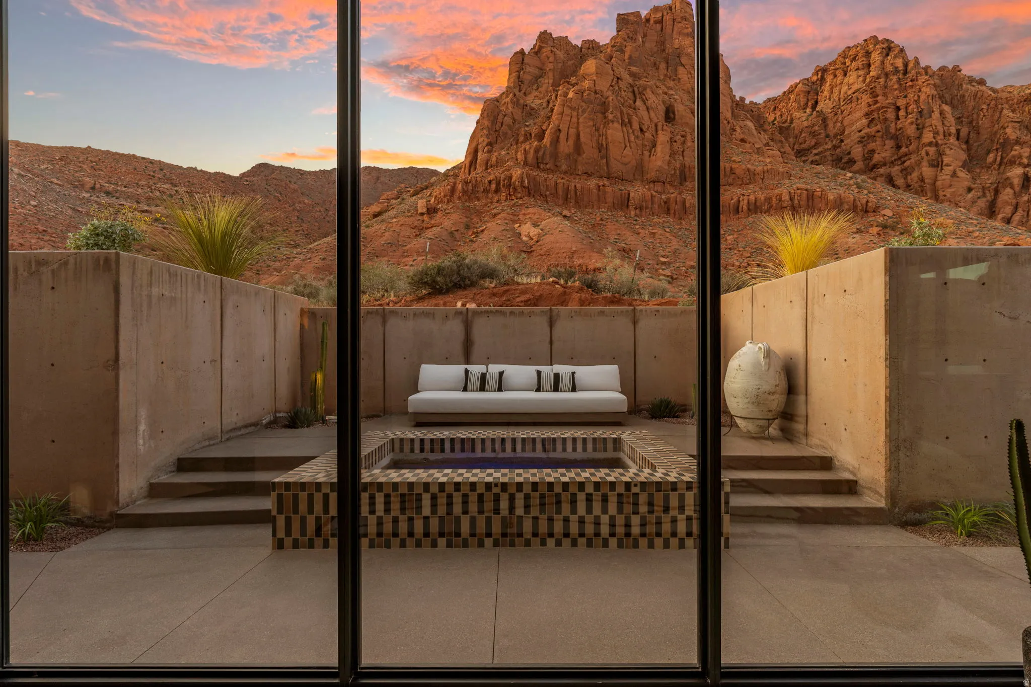 Expansive glass doors opening to an outdoor patio with a tiled water feature, concrete retaining walls, and tiered hardscaping, all set against a backdrop of rugged red rock mountains