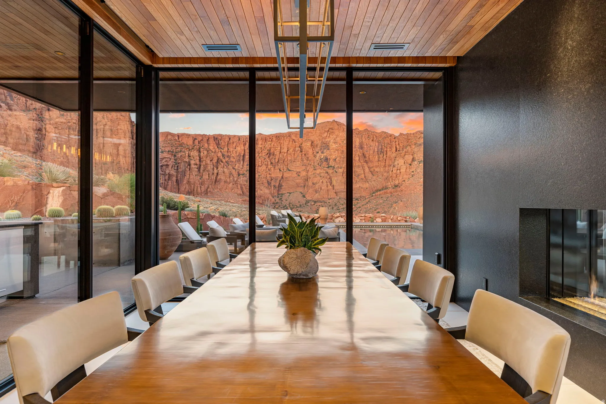 Floor-to-ceiling windows, wood-paneled ceiling, linear fireplace with a dark-textured surround, and a long dining surface