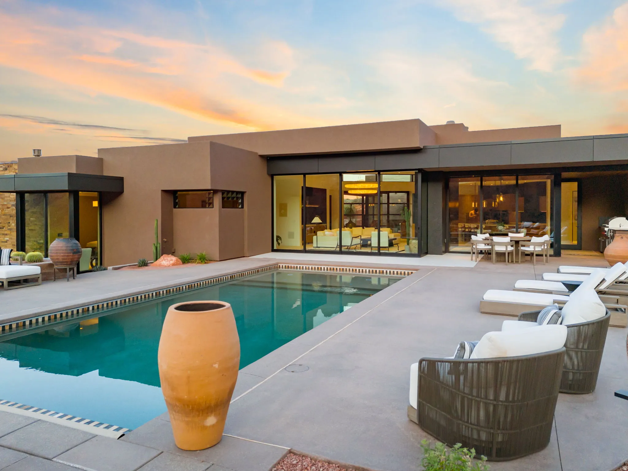 Modern exterior architecture featuring a flat roofline, expansive concrete patio, rectangular swimming pool, large sliding glass doors, and desert landscaping