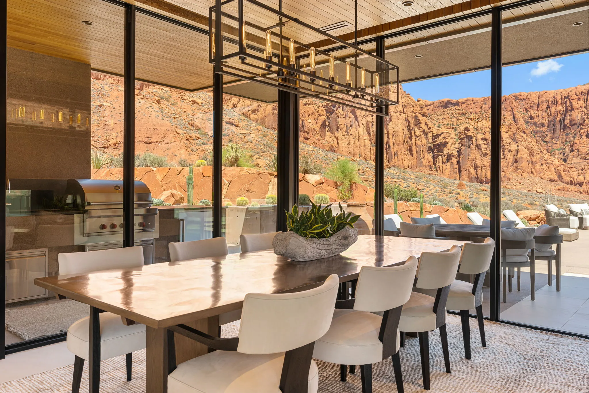 Floor-to-ceiling glass panels, wood-finish ceiling, linear chandelier, built-in outdoor grill station, and red rock formations