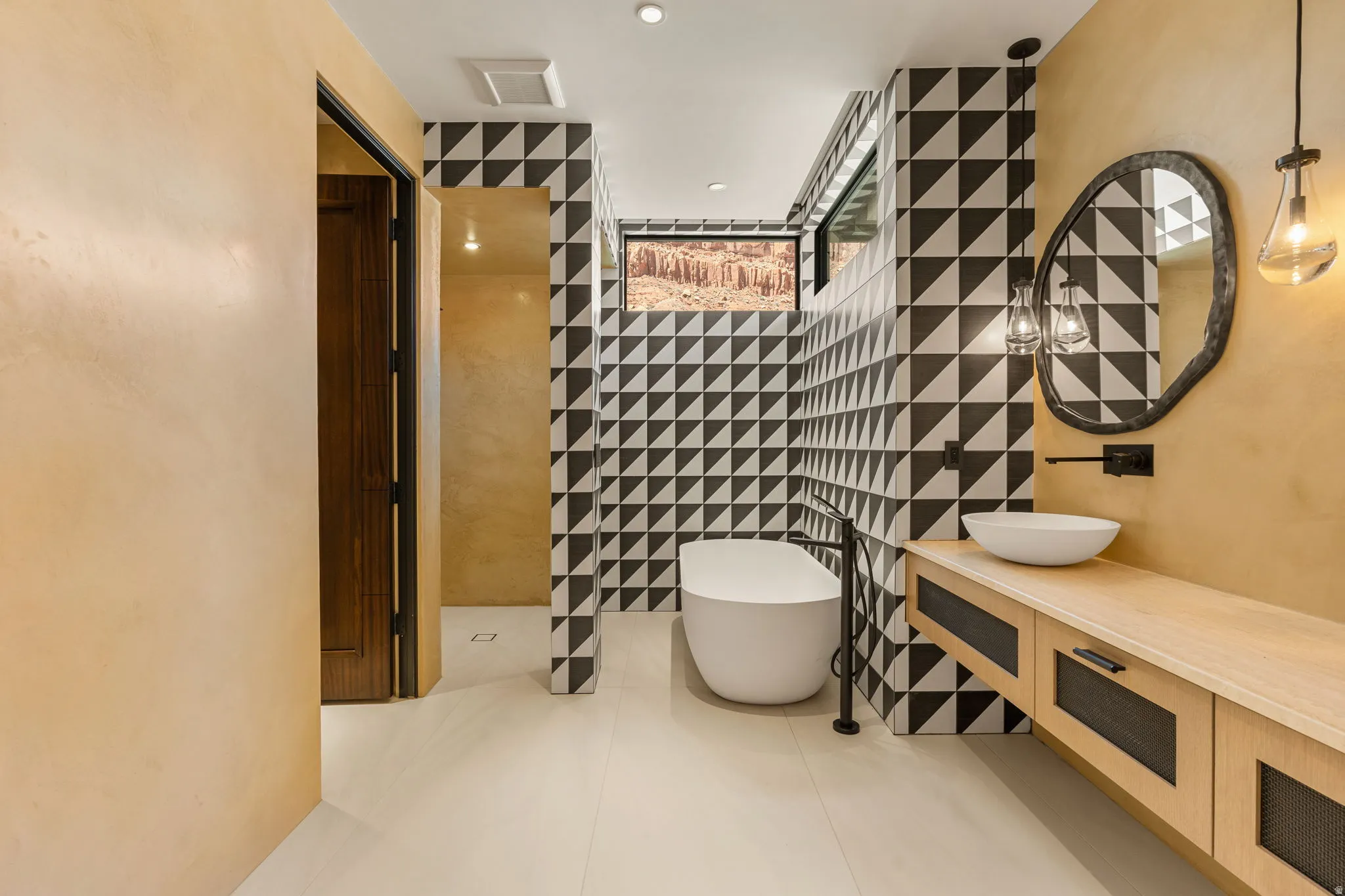 Freestanding soaking tub, black and white geometric wall tiling, floating vanity with vessel sink, wall-mounted faucet, and custom mirror