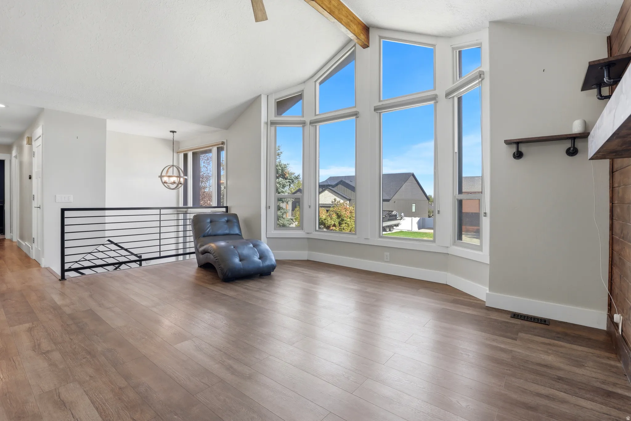 Vaulted ceiling with exposed wood beam, large bay window array, wood-finish flooring, black horizontal bar railing, and a rustic wood mantel with integrated shelving