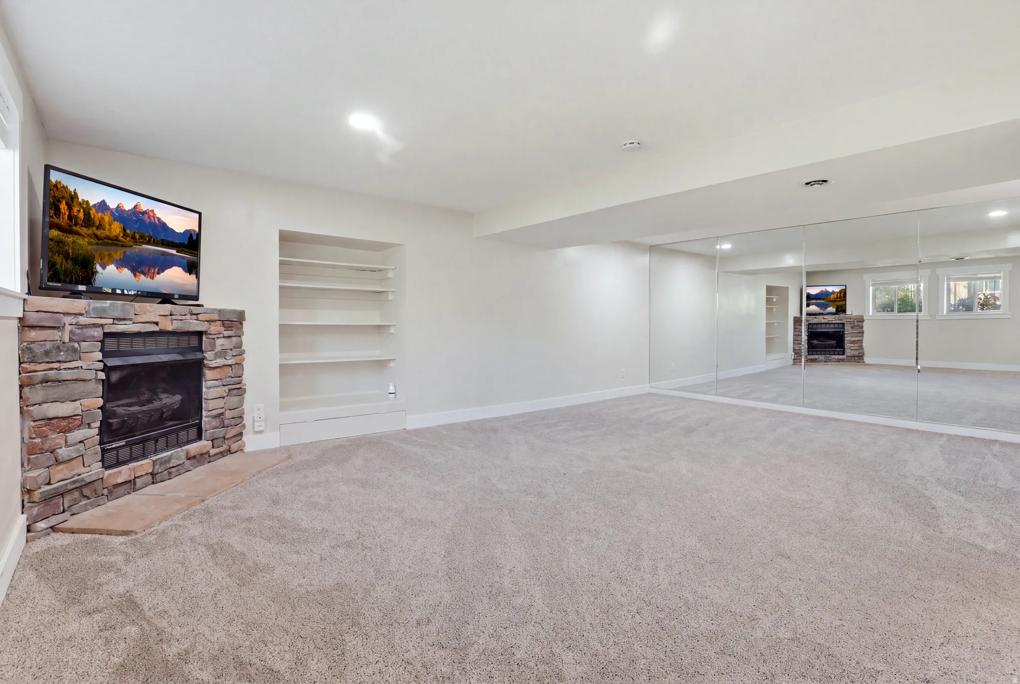 Stone-clad fireplace with a hearth, built-in shelving, mirrored wall, recessed lighting, and light-toned carpeting