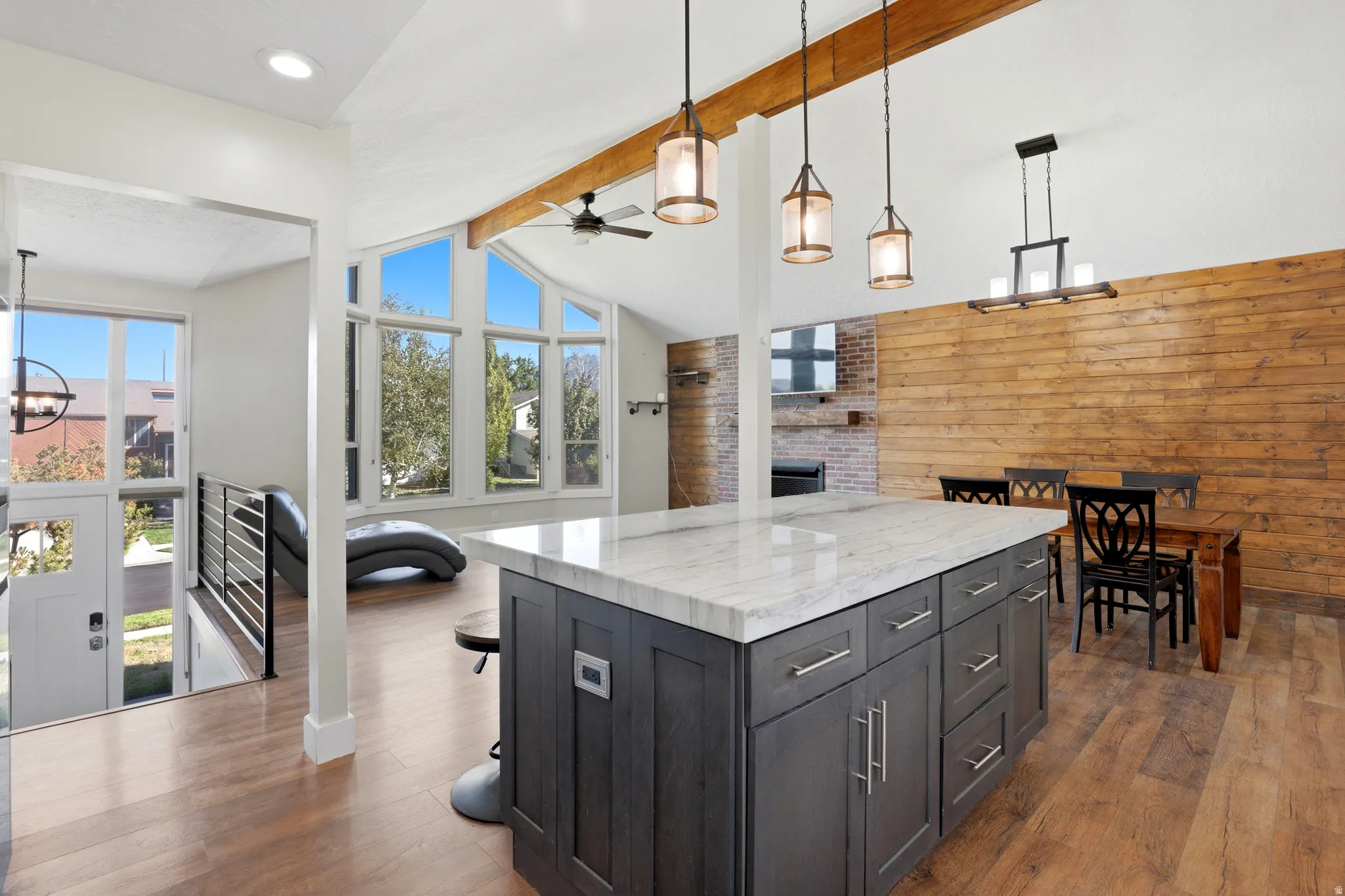 Vaulted ceiling with wood beam accents, large island with stone countertop, wood-finish flooring, accent wall with horizontal wood paneling, and a brick fireplace