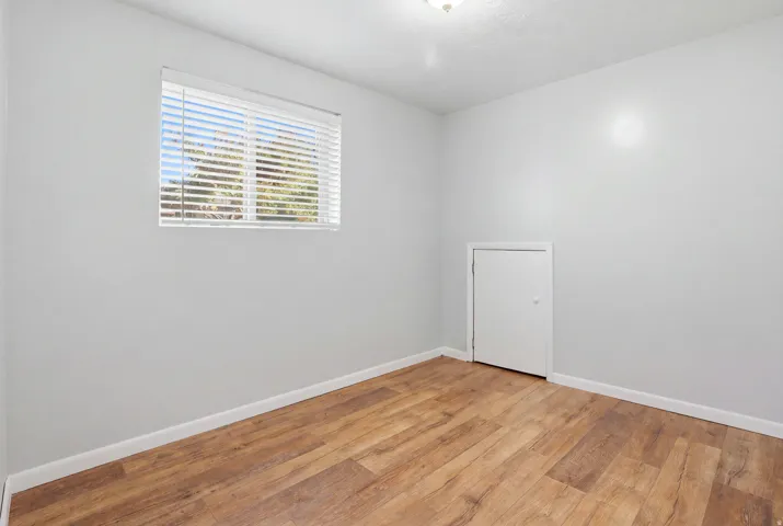 Wood-finish flooring with white baseboards, a single window with blinds, a white door, and a flush mount ceiling light