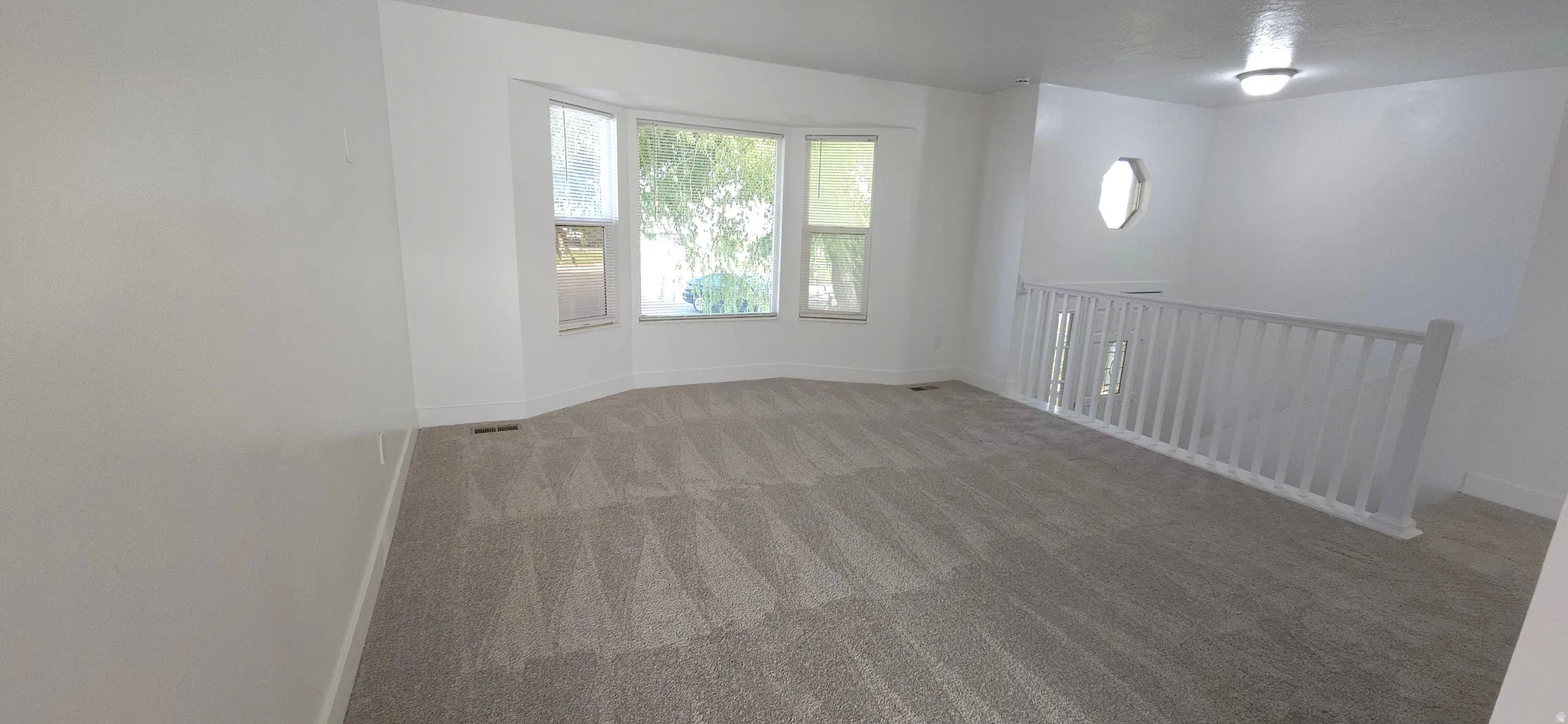 Spacious room featuring a bay window, neutral carpeting, white balustrade, and an upper window