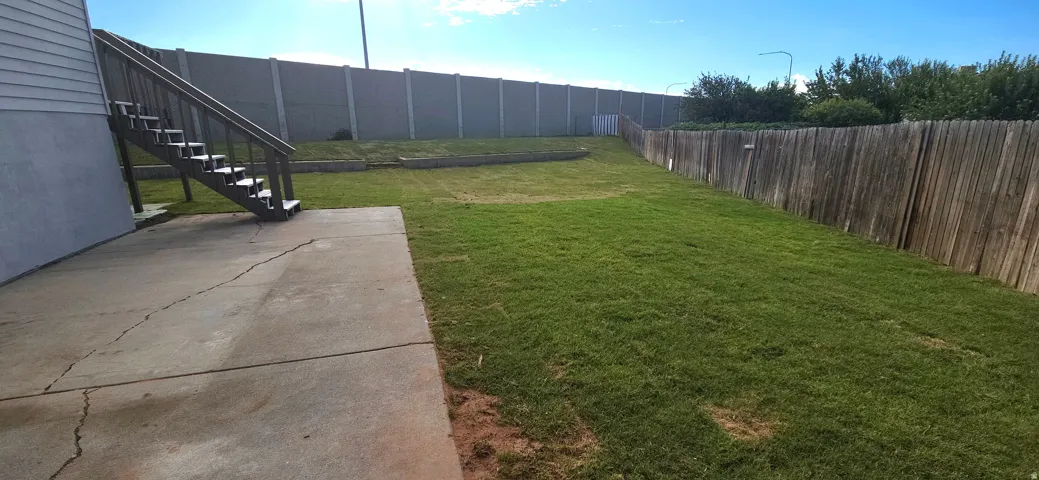 Expansive grassy yard with concrete patio, wood fencing, and a concrete privacy wall