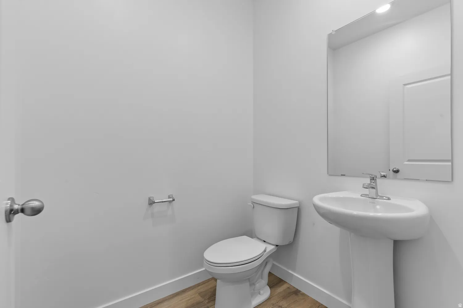 Pedestal sink with chrome faucet, wall-mounted mirror, white toilet, and wood-finish flooring