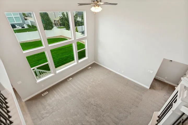 Two-story ceiling, large multi-pane windows, neutral-toned carpet flooring, ceiling fan with integrated lighting, and white baseboards
