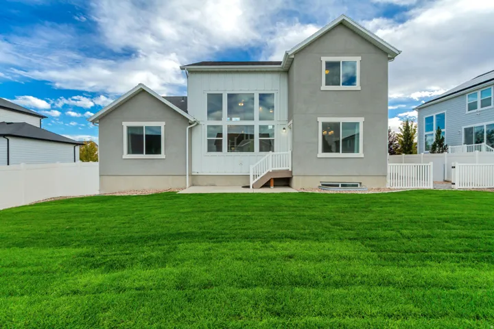 Expansive green lawn with a stucco and siding exterior, multiple large windows, a white privacy fence, and a concrete patio