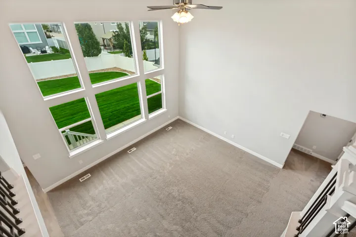 Two-story ceiling, large multi-pane windows, neutral-toned carpet flooring, ceiling fan with integrated lighting, and white baseboards