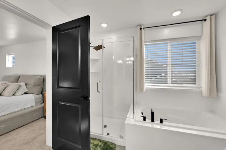 Recessed lighting, a dark paneled door, glass-enclosed shower, a built-in soaking tub, and a window with horizontal blinds