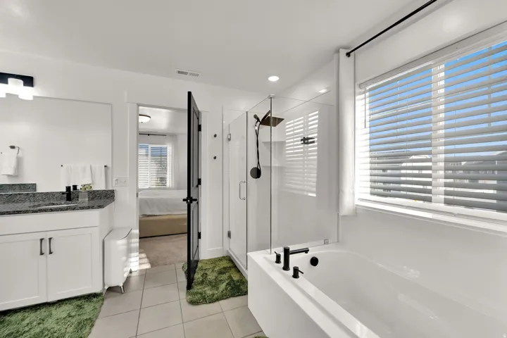 Tiled flooring featuring a built-in soaking tub with matte black fixtures, a glass-enclosed shower, a vanity with a dark countertop and white cabinetry, and a window with blinds