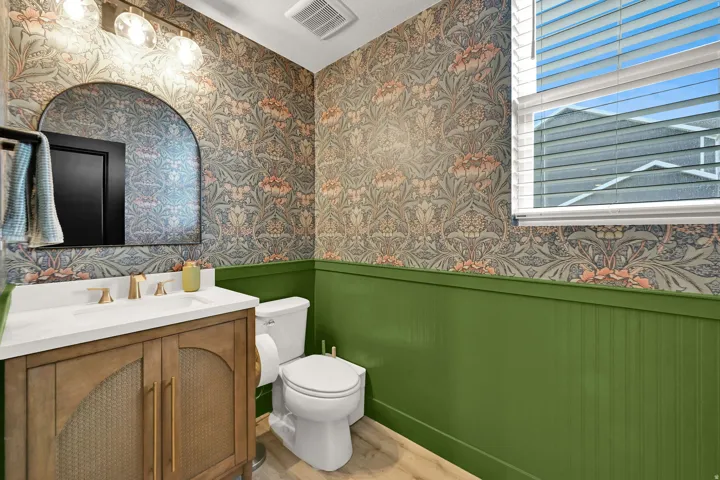 Arched vanity mirror with a three-light fixture, patterned wallpaper, green beadboard wainscoting, and wood-finish flooring