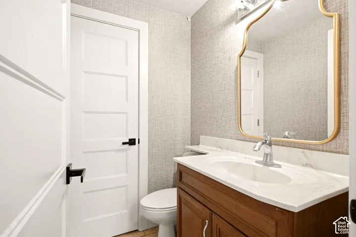 Neutral-toned textured wallcovering, vanity with a stone-top surface and undermount sink, decorative gold-tone framed mirror, and matte black door hardware