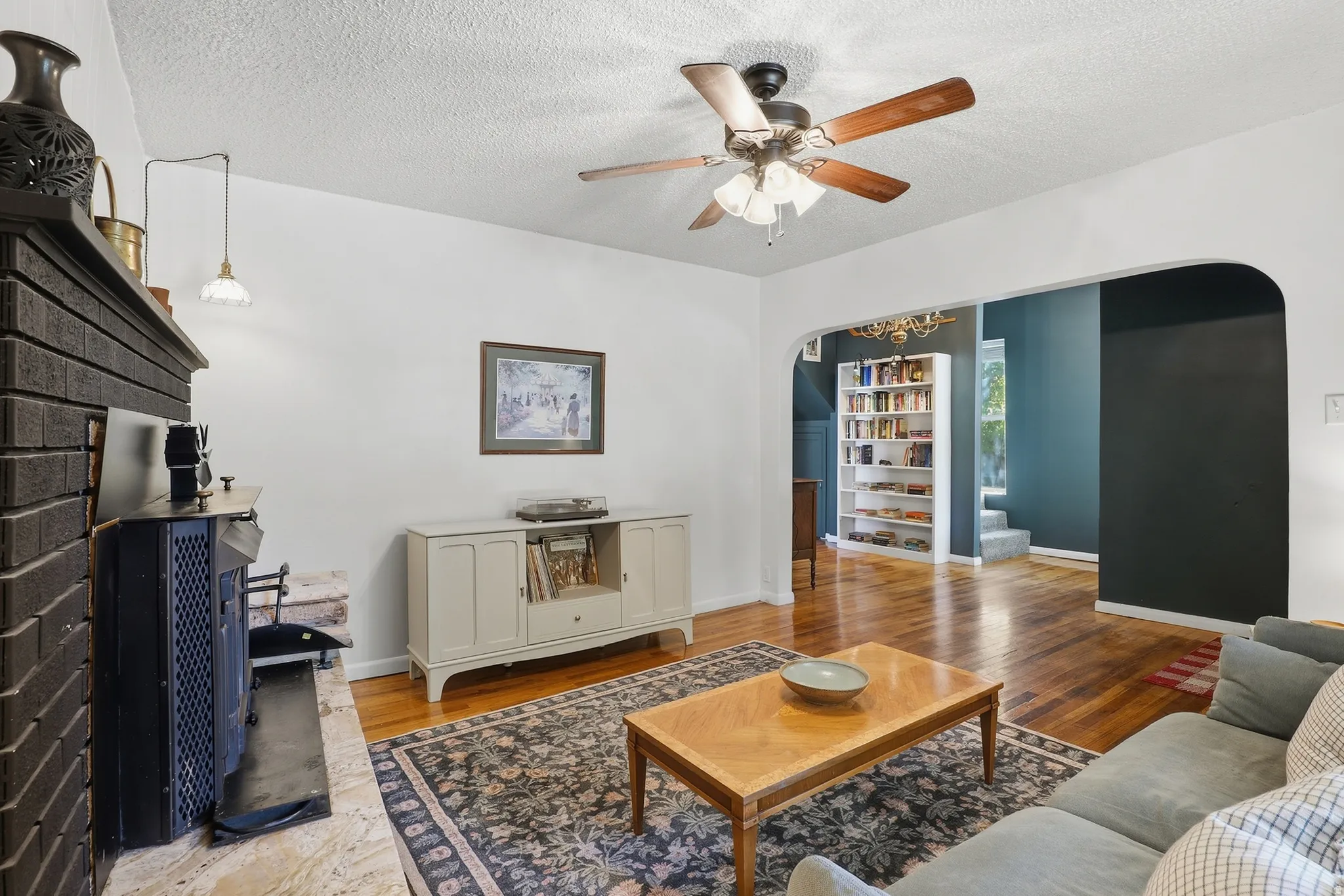 Arched doorways, hardwood flooring, fireplace with a brick surround, ceiling fan, and built-in shelving