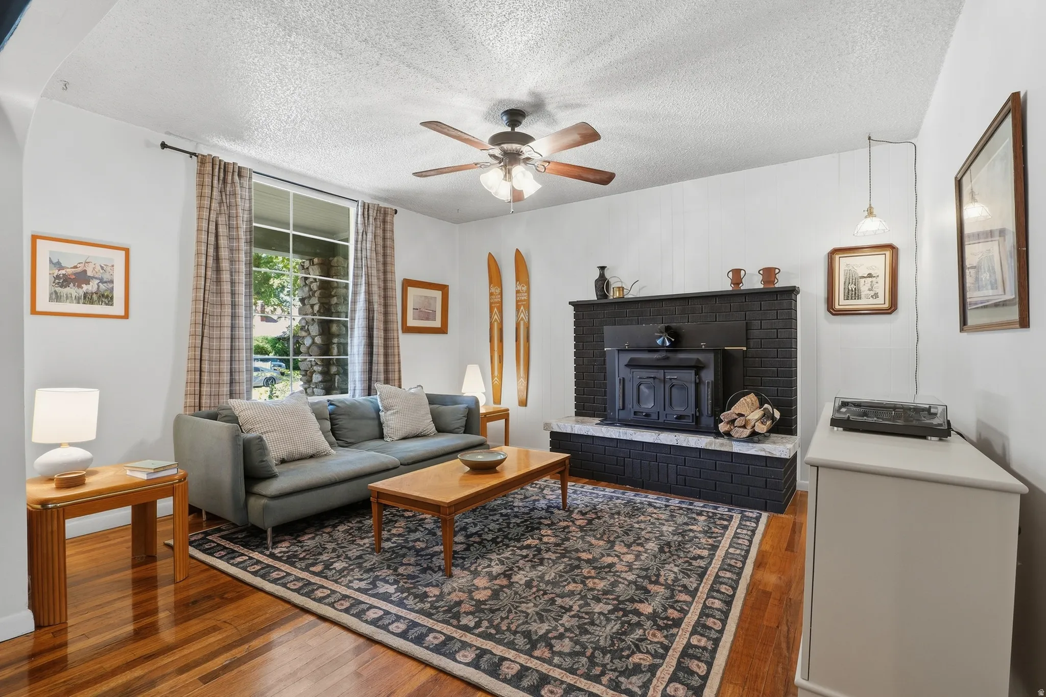 Hardwood flooring, ceiling fan, large window, black brick fireplace with a wood-burning stove, and a stone hearth