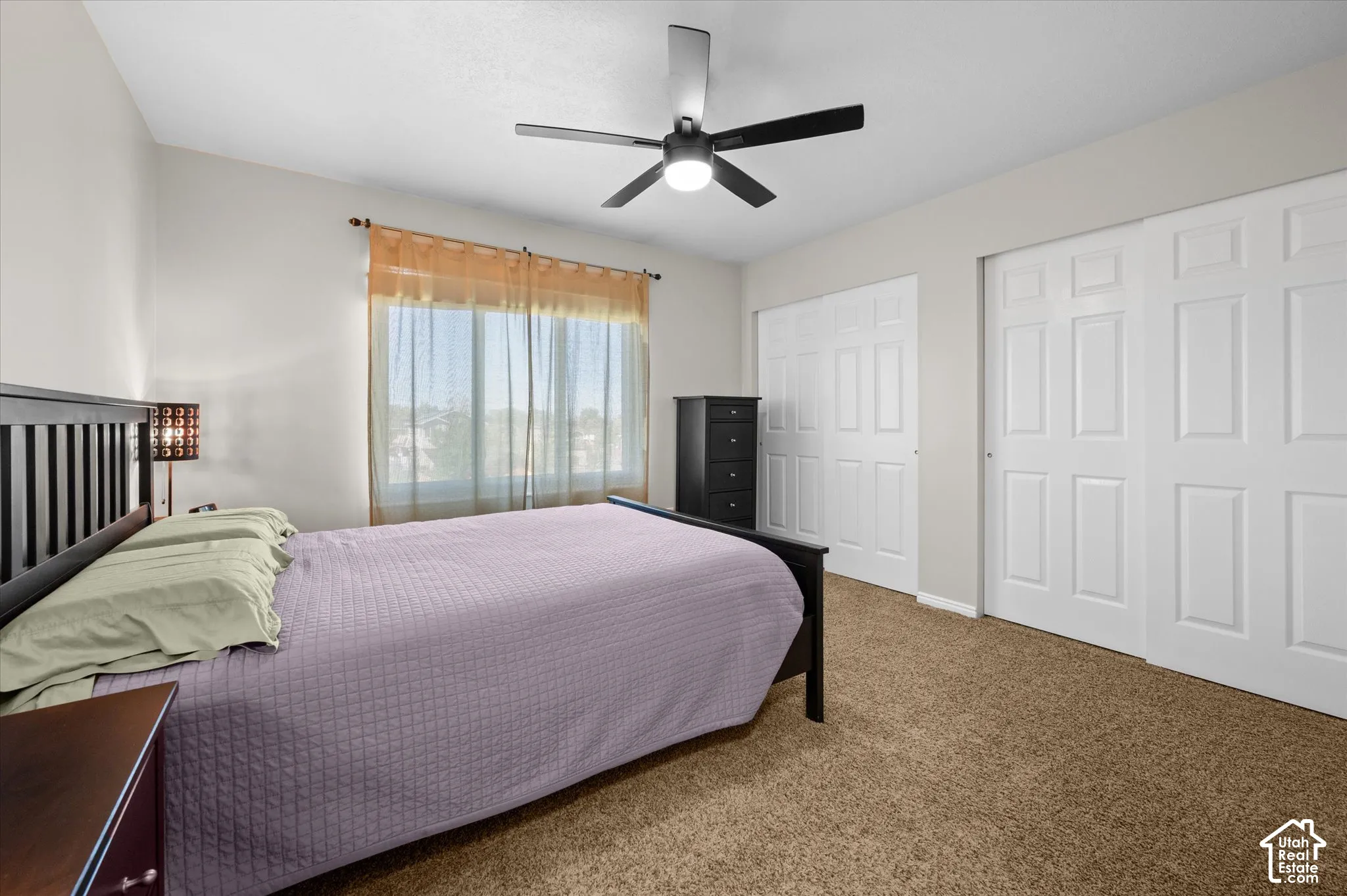 Carpeted room featuring a ceiling fan with integrated lighting, a window with sheer curtains, and two sets of bi-fold closet doors