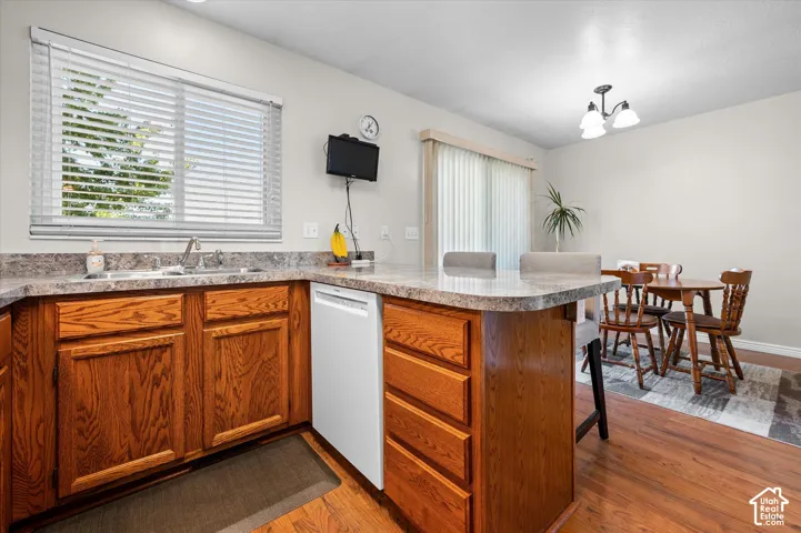 Kitchen featuring wood cabinetry, a double basin sink, a white dishwasher, light-toned countertops, and wood-finish flooring