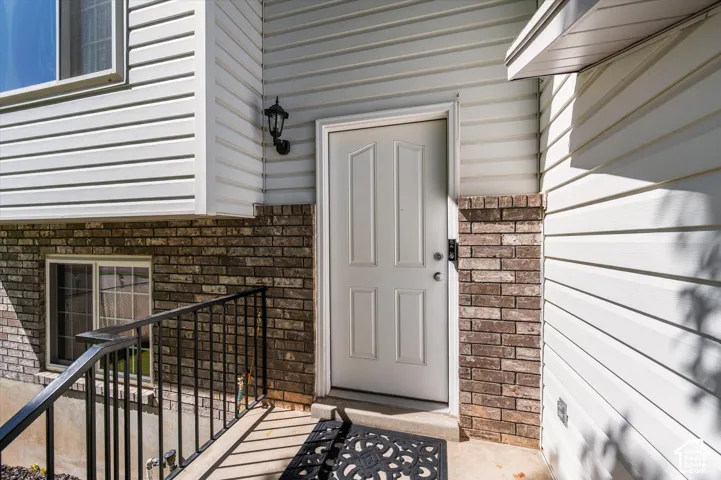 White paneled entry door with an overhead light fixture, brick detailing, white horizontal siding, and a black metal handrail with box window
