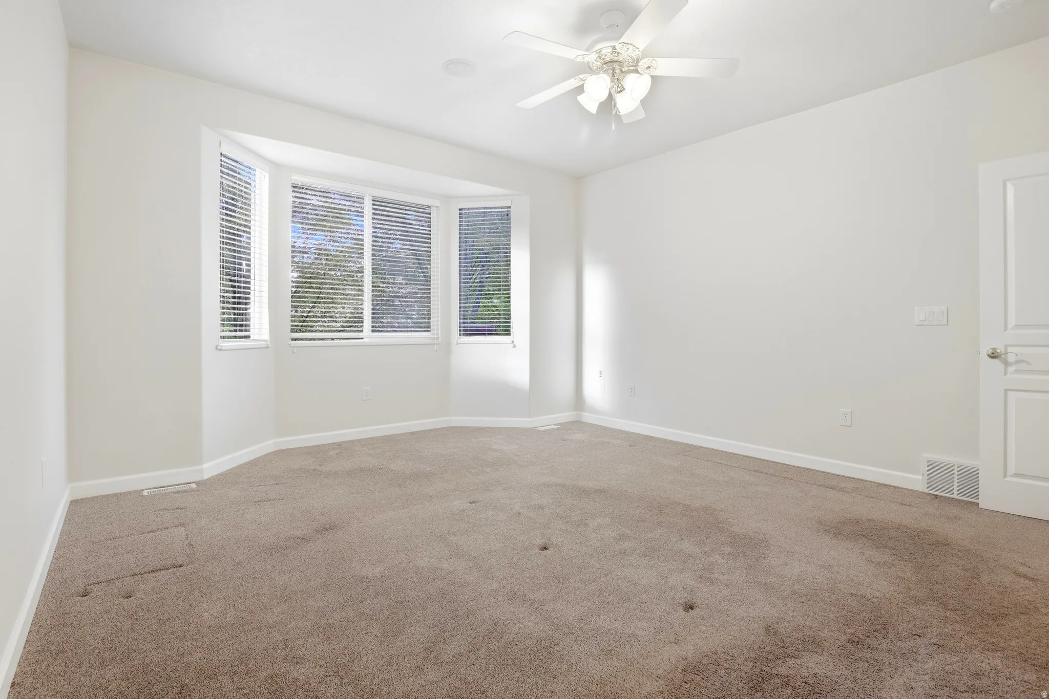 Bay window with horizontal blinds, ceiling fan, white paneled door, and neutral carpeting