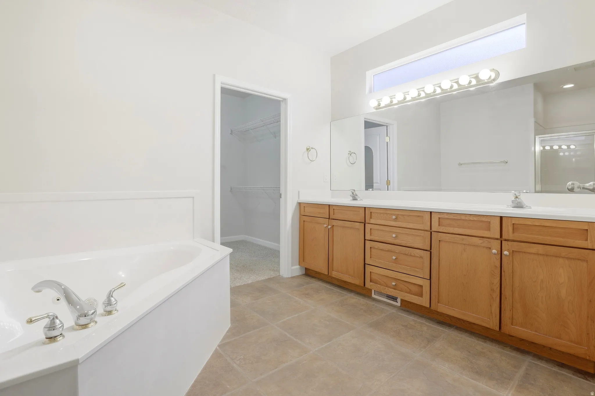 Soaking tub with chrome fixtures, dual vanity with wood cabinetry, and a large mirror with integrated lighting