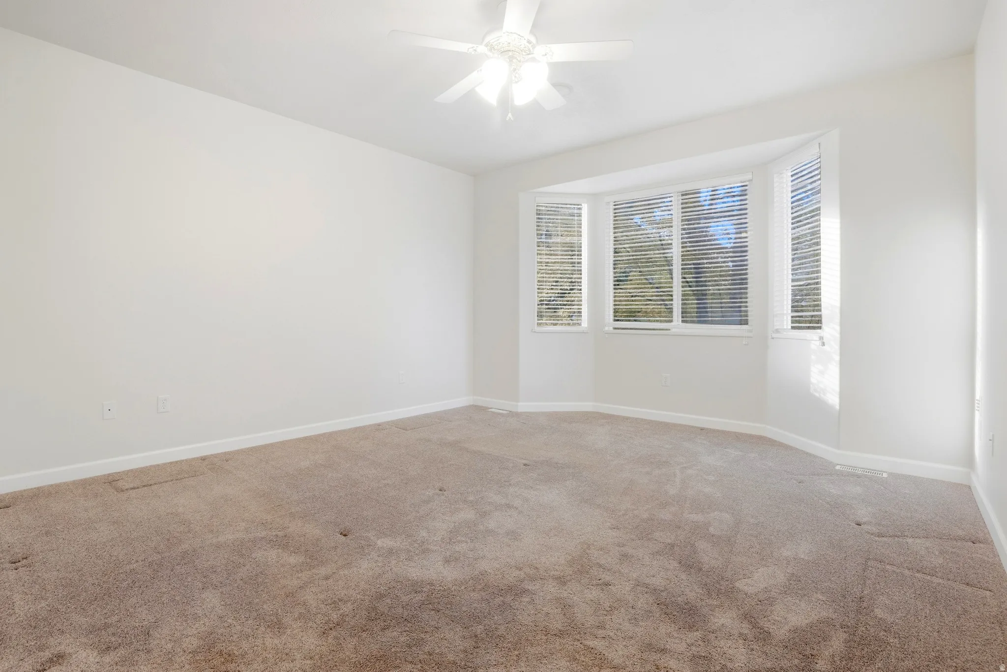 Bay window with horizontal blinds, neutral-toned carpeting, white walls, and a ceiling fan with integrated lighting