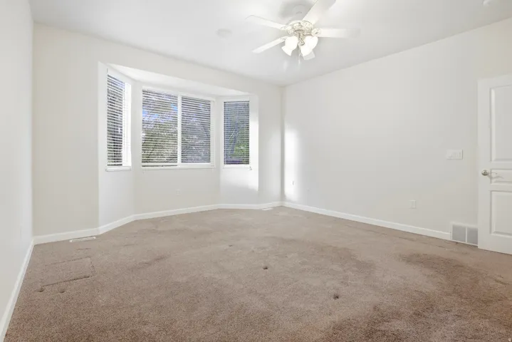 Bay window with horizontal blinds, ceiling fan, white paneled door, and neutral carpeting