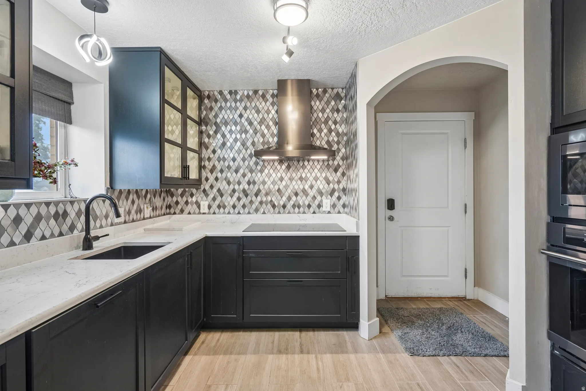 Kitchen featuring dark cabinetry, light countertops, mosaic tile backsplash, wood-finish flooring, and a stainless steel range hood