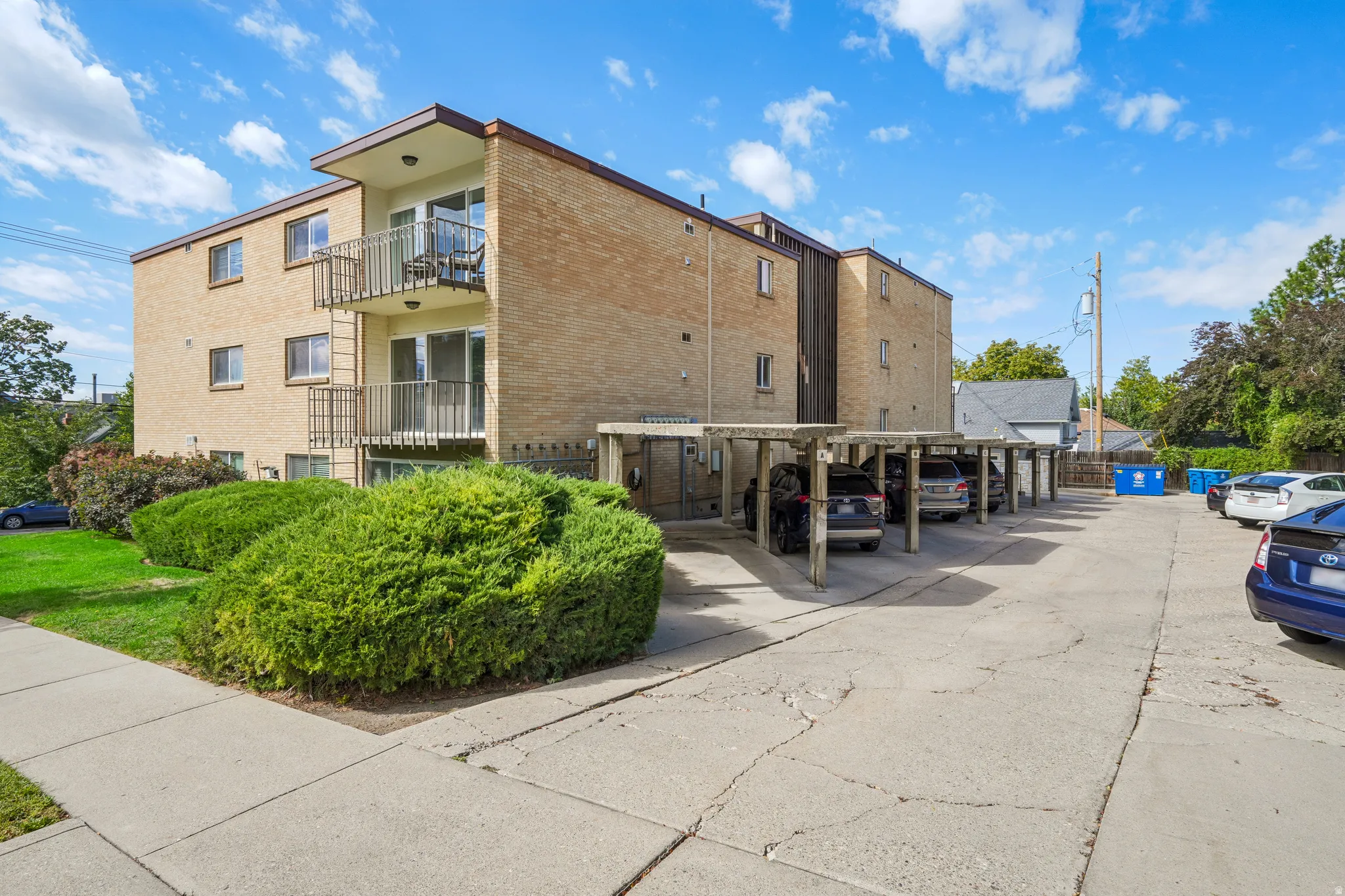 Brick exterior, balconies with metal railings, covered parking area, paved parking lot, and mature landscaping