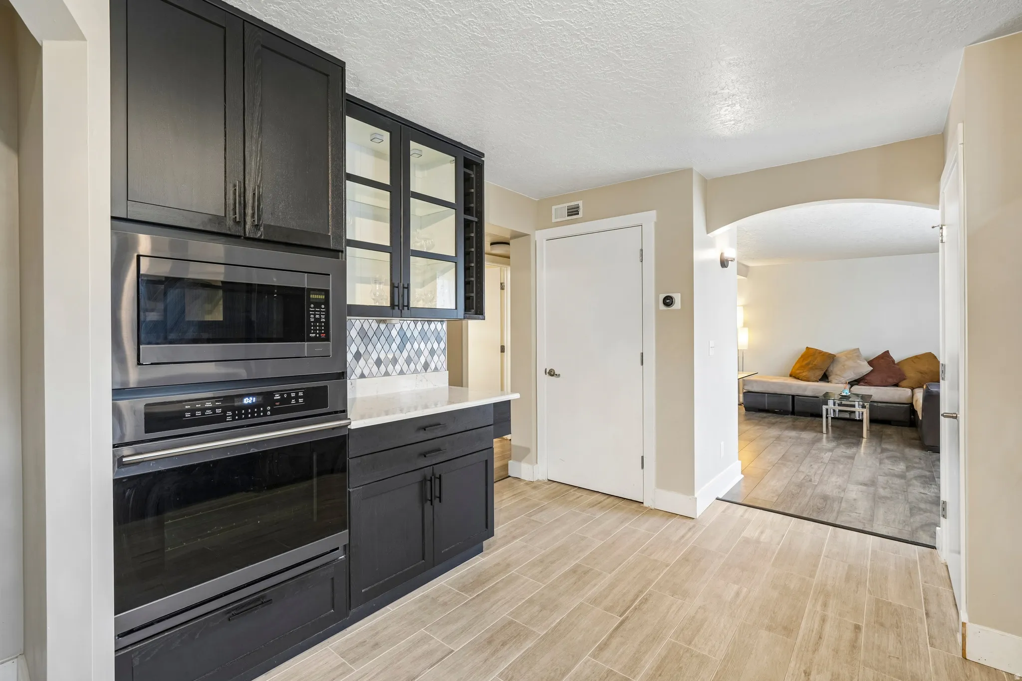 Built-in stainless steel microwave and oven, dark cabinetry with glass-front accents, mosaic tile backsplash, light wood-finish flooring, and an arched doorway