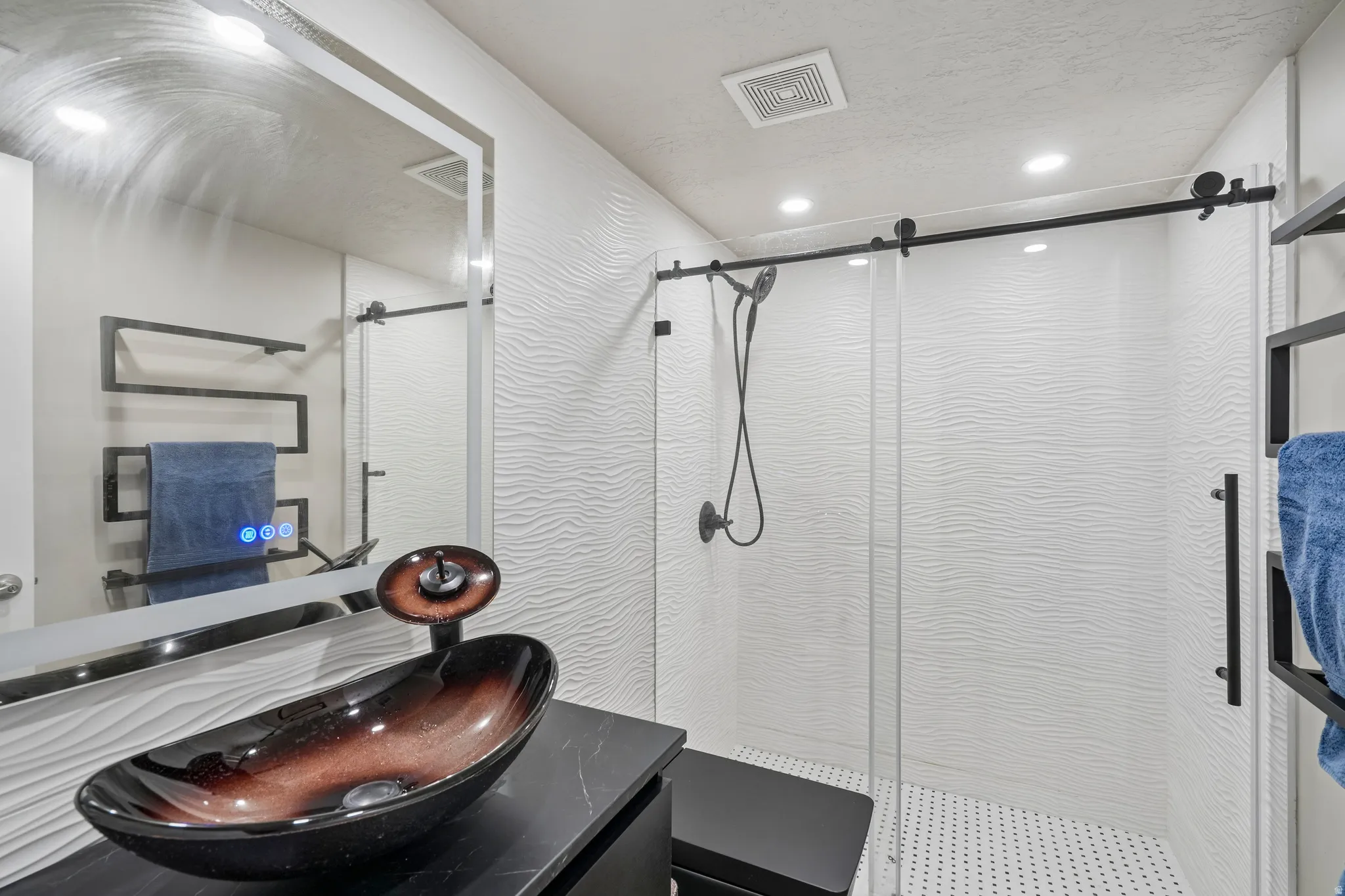 Recessed lighting illuminates the modern vanity with a vessel sink and black countertop, complemented by a glass-enclosed shower with a barn door style entry and textured wall tile