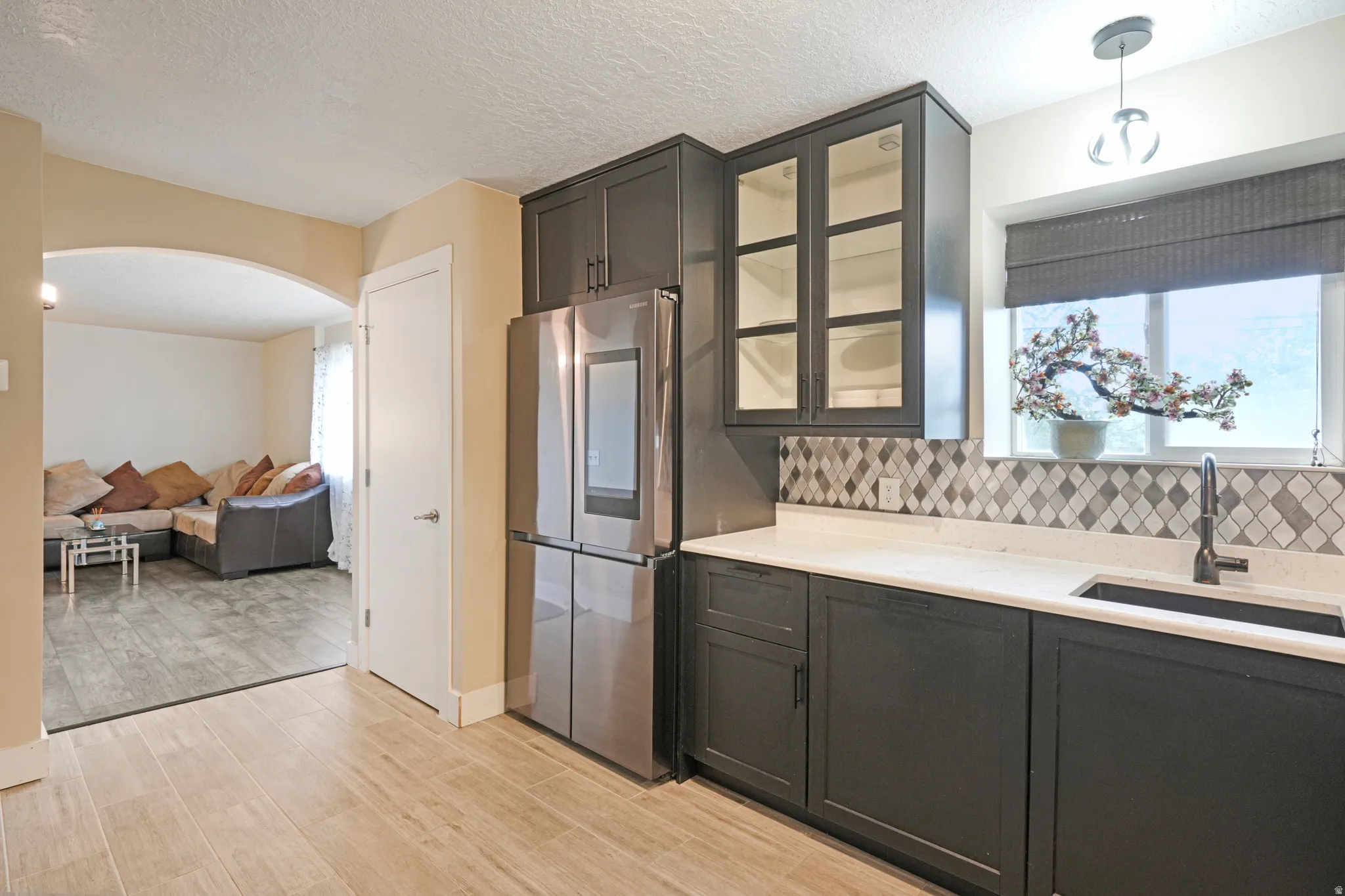 Wood-finish flooring, dark cabinetry with glass-front uppers, stainless steel refrigerator, white countertops, and a decorative tile backsplash