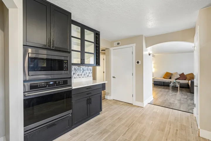 Built-in stainless steel microwave and oven, dark cabinetry with glass-front accents, mosaic tile backsplash, light wood-finish flooring, and an arched doorway