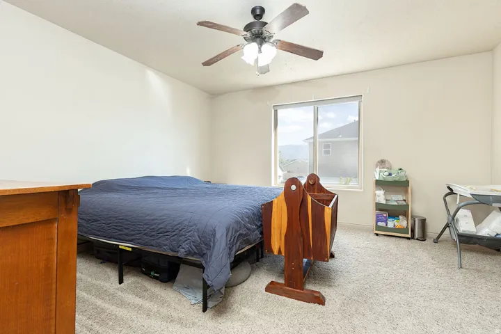 Light-colored wall finishes, carpet flooring, a window with a roller shade, and a ceiling fan with integrated lighting