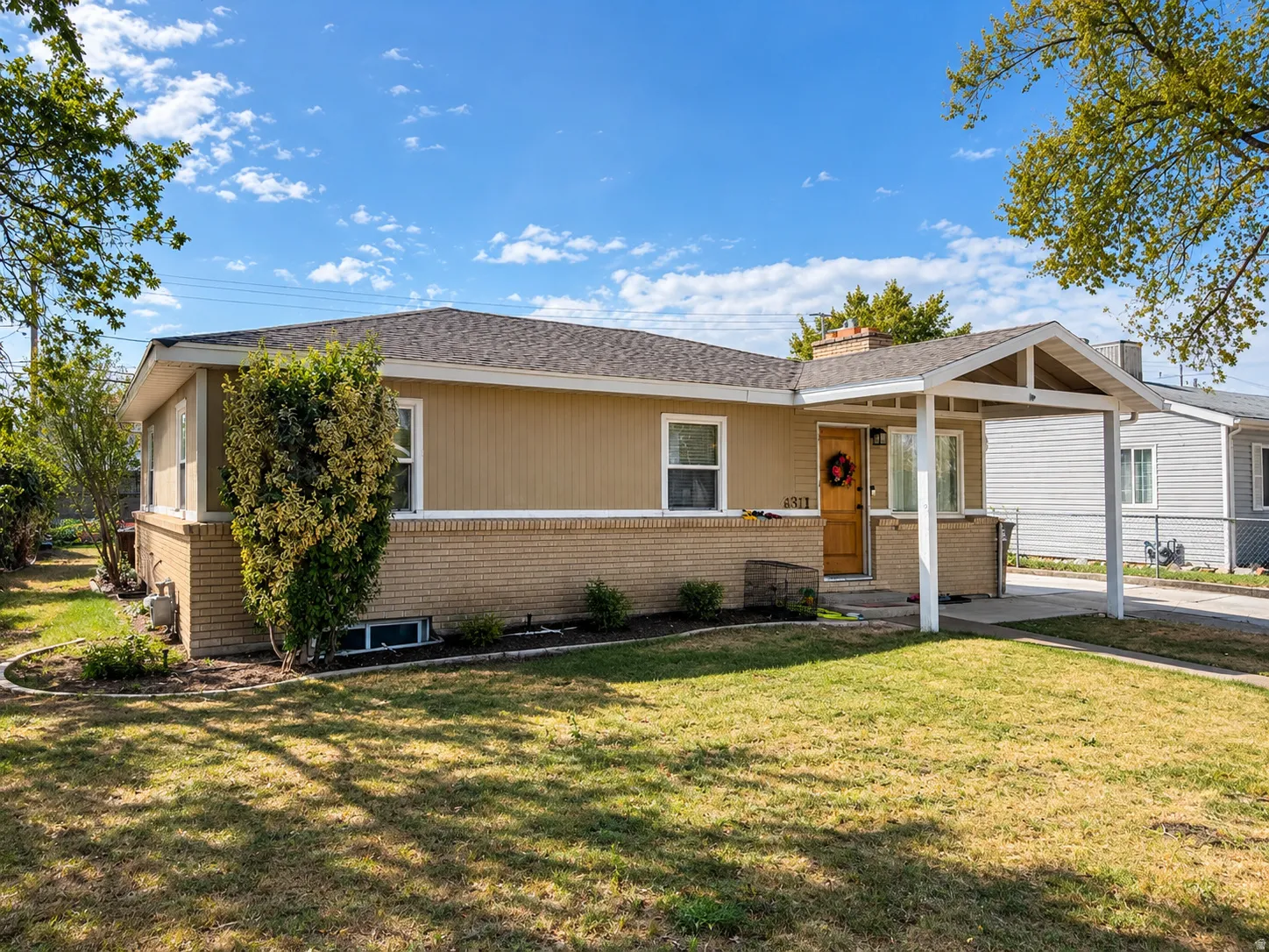 Tan siding exterior with a brick lower facade, gabled carport, and mature front landscaping